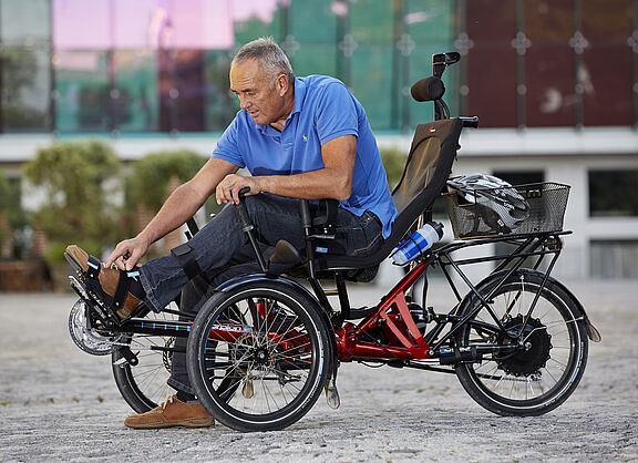 Ein Mann in blauem Poloshirt sitzt auf einem roten dreirädrigen Elektrofahrrad mit schwarzer Rückenlehne und Korb. Das Spezialrad steht auf einem gepflasterten Platz vor einem modernen Gebäude mit grünen Fenstern.