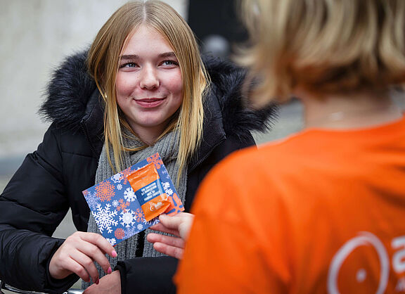 Eine lächelnde junge Frau in Winterjacke erhält von einer ADFC-Person eine blaue Karte mit Schneeflocken-Motiv und einen Zimtstern.